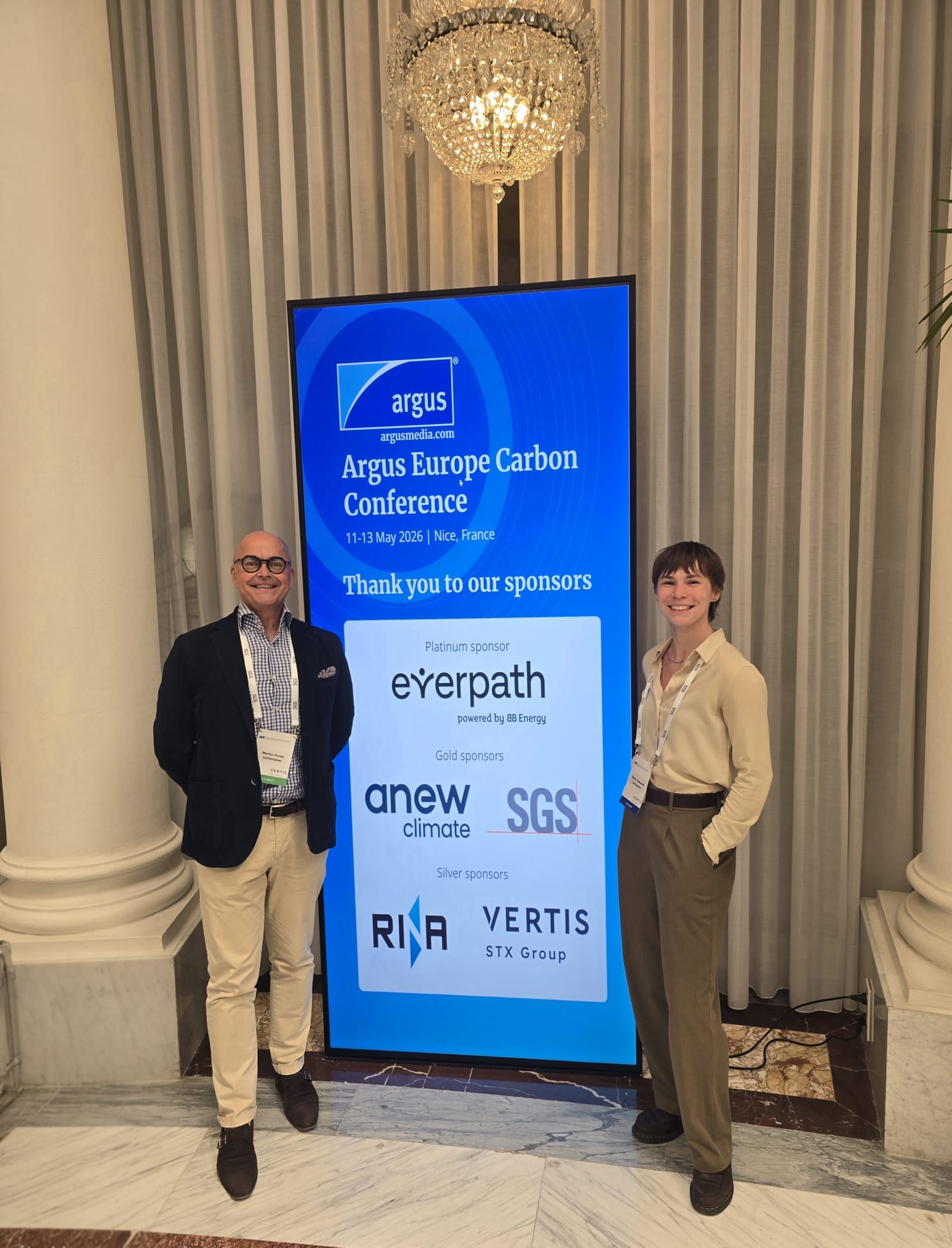 Carbonaires team members Martyn Porter and Marika Niekowal at the Argus Europe Carbon Conference 2026 in Nice, standing on either side of a large vertical digital screen showing the Argus Europe Carbon Conference branding and sponsor logos