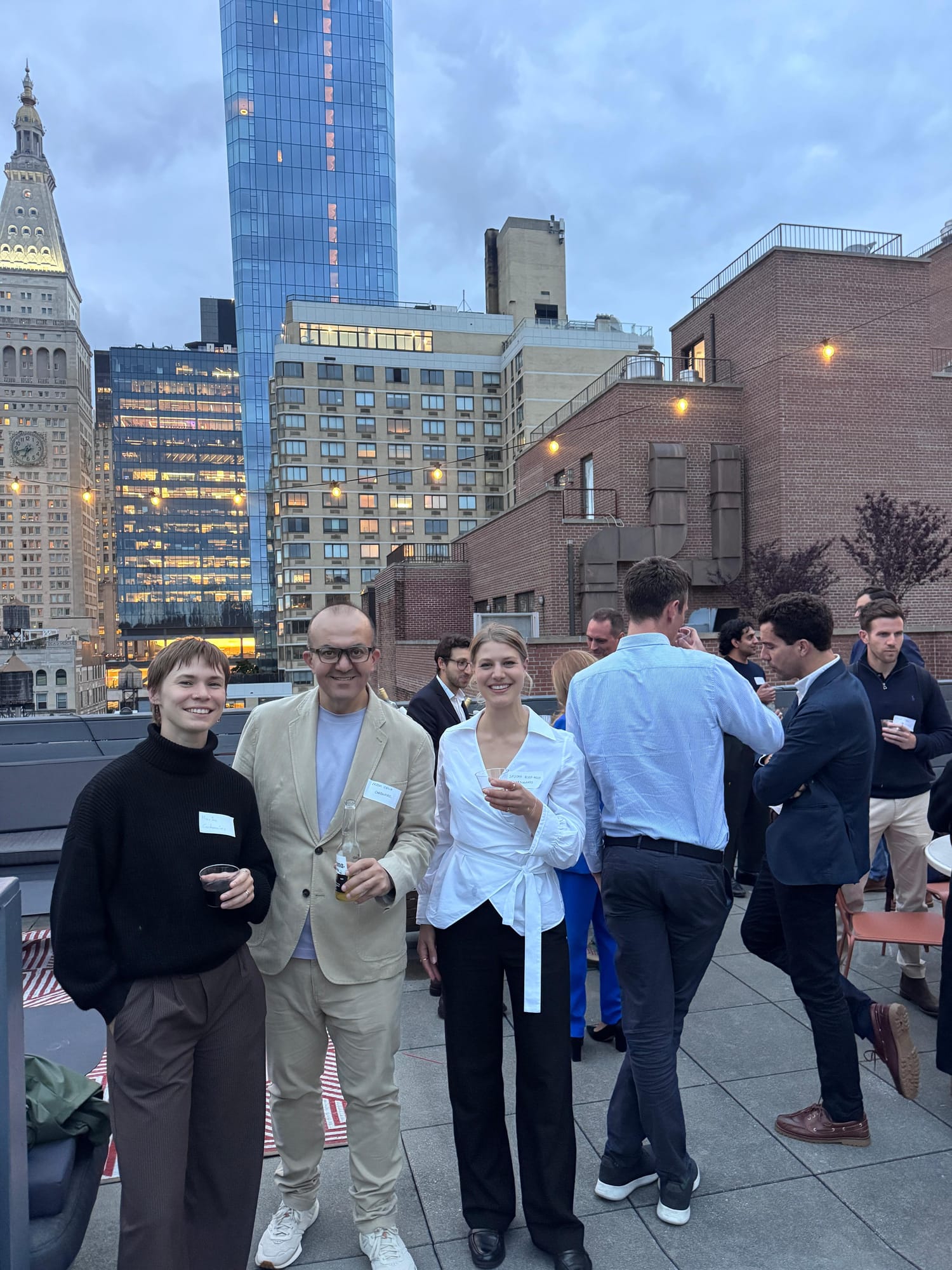 Roundtable participants talking over drinks on a rooftop terrace in New York at dusk, with lit high-rise buildings behind them