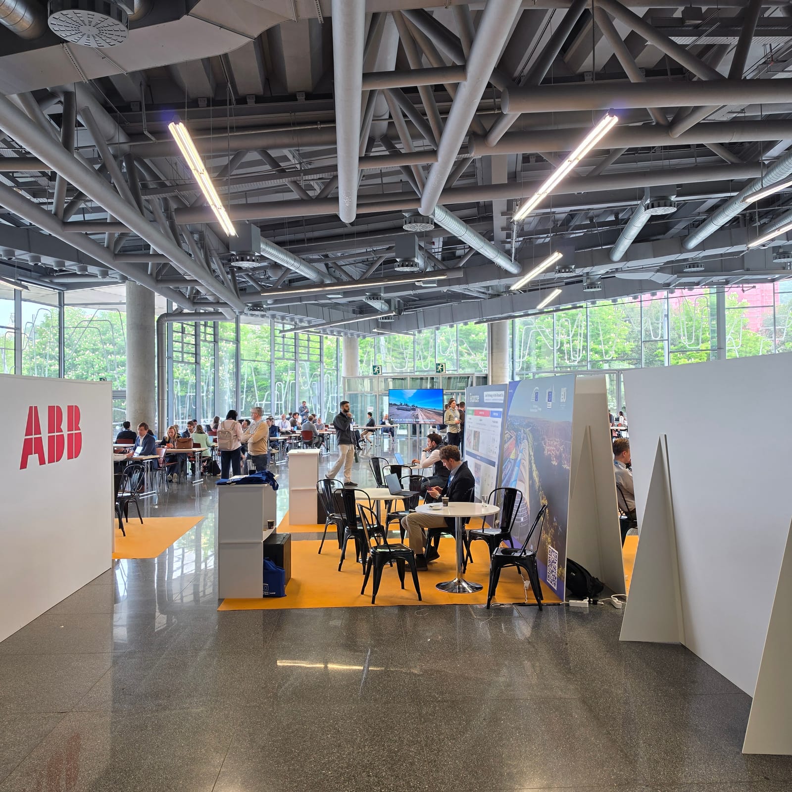Energy Tech Summit 2026 networking mezzanine in Bilbao with delegates seated and standing at high tables, ABB sponsor signage in the foreground and the venue’s exposed steel ceiling structure overhead