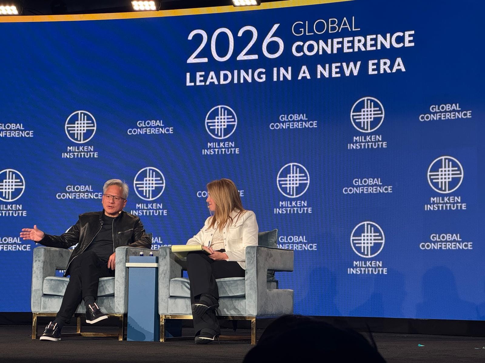 NVIDIA CEO Jensen Huang in a fireside chat on stage at the Milken Institute Global Conference 2026, against a blue Milken Institute backdrop reading "2026 Global Conference: Leading in a New Era"