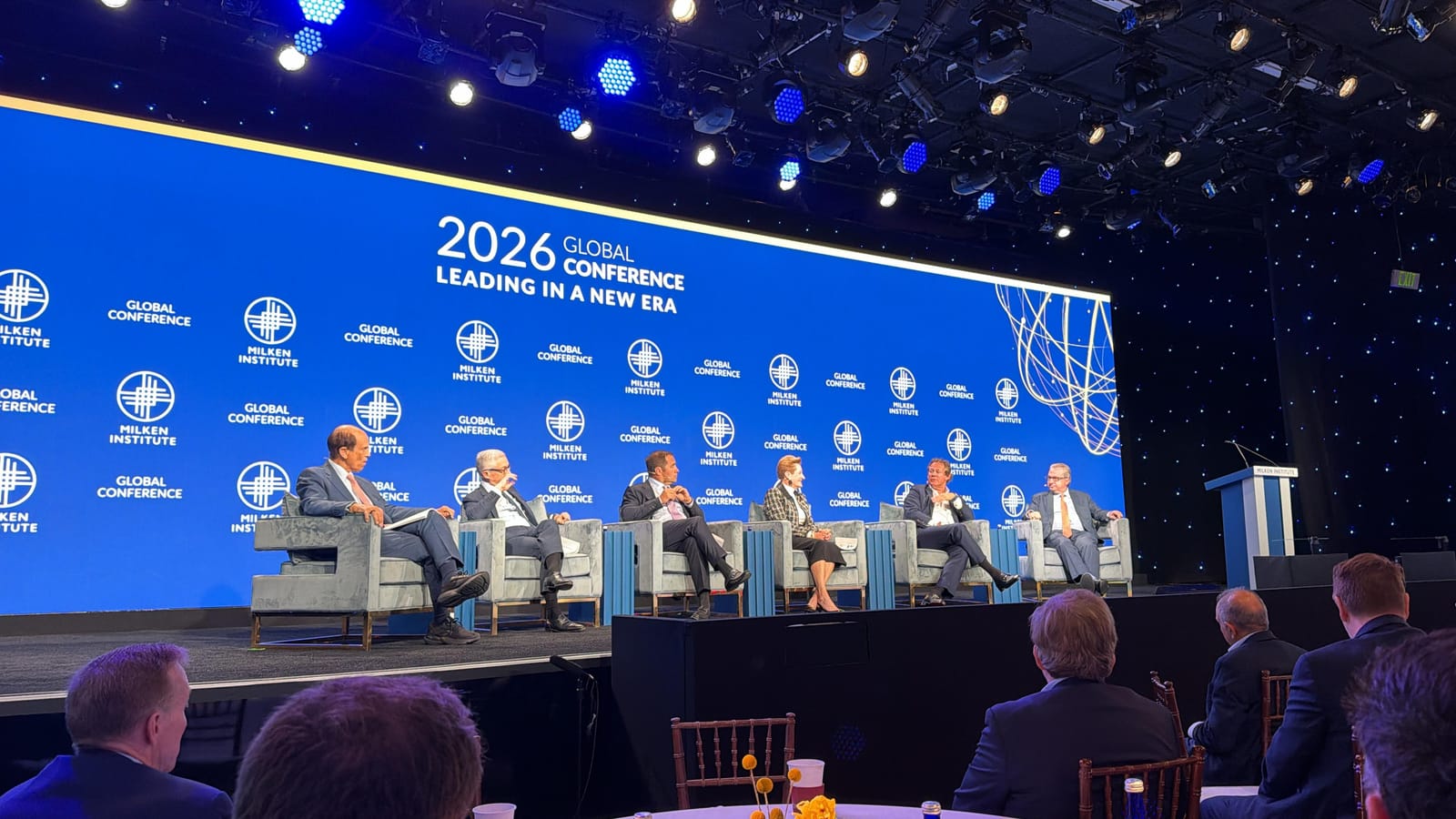 A six-person panel discussion on stage at the Milken Institute Global Conference 2026, with delegates seated in the audience and a large blue Milken Institute backdrop reading "2026 Global Conference: Leading in a New Era"
